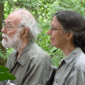 An elderly man with white hair and beard, wearing glasses and a loose-fitting khaki top, along with a dark-haired woman with her hair tied back, glasses, and a similar khaki top, both in a Costa Rican rainforest.