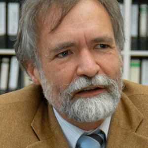 Portrait of an old man with greying hair and beard, wearing a tan suit over a white shirt and powder blue tie, in front of a bookcase.