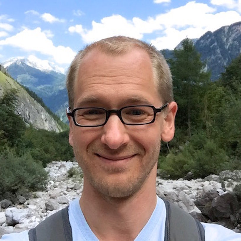 Dr. Florian Leese, a middle-aged white man wearing glasses, a blue shirt, and a vest, in front of a mountainous landscape.