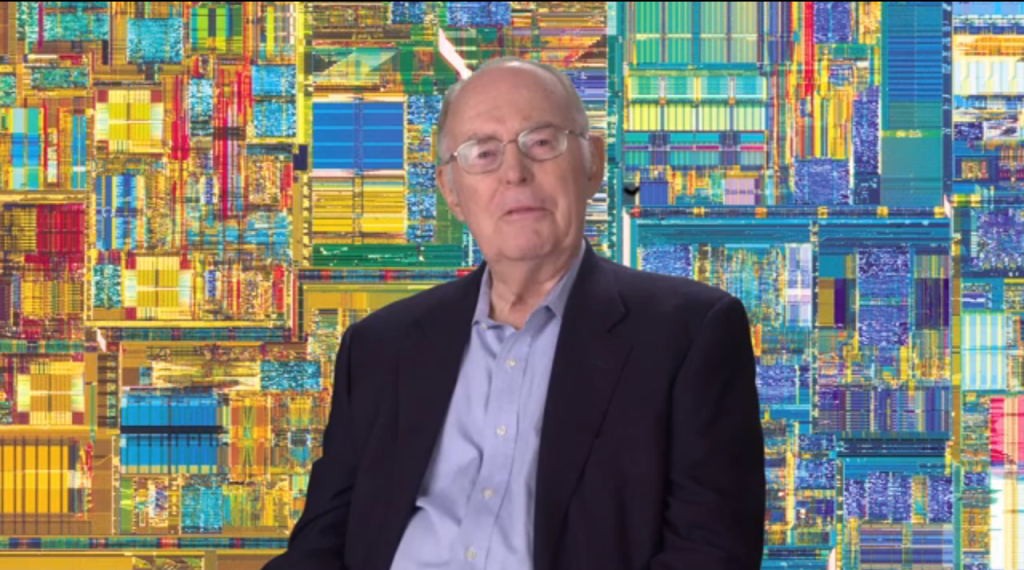 Gordon Moore wearing a suit jacket and shirt and sitting in front of a microchip themed backdrop.